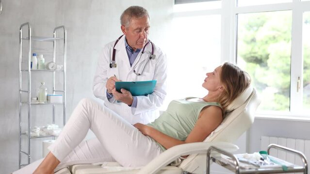 Old man physician making notes while discussing treatment procedures with female patient lying nearby