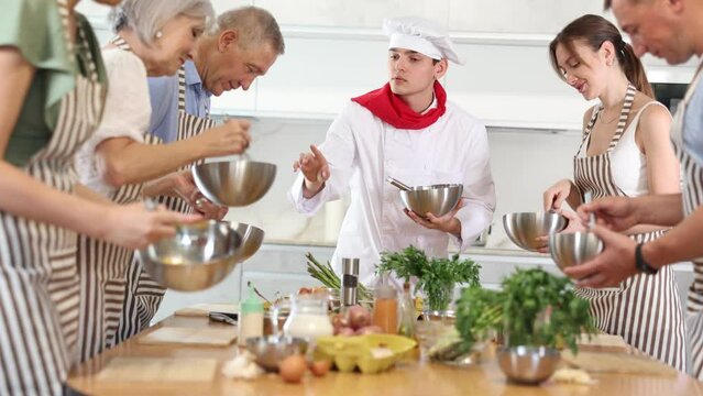 Smiling young man cook conducting culinary classes for learners standing around kitchen table with whisk and bowl in hands
