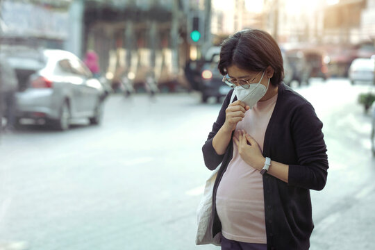 Asian pregnant woman wearing PM2.5 dust mask and coughing on the street. wearing mask protect against pollution, anti smog and viruses, air pollution, and headache suffocating. City air pollution.