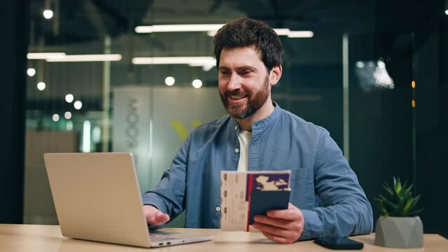 Satisfied entrepreneur using computer while resting hand with cruise admission ticket in passport on desktop with succulent. Happy man checking living conditions in cabin on ship on company website.