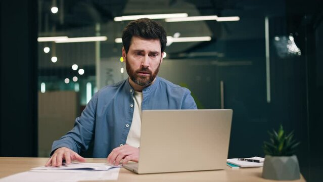 Portrait of caucasian man checking business documentation and typing on laptop while sitting indoor. Confused accountant struggling with filling out tax return in printed and electronic forms.