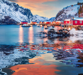 Red wooden houses of Nusfjord town and snowy peaks reflected in the calm waters of Vestfjorden fjord, Norway, Europe. Amazing sunset on Lofoten Island archipelago. Life over polar circle. - obrazy, fototapety, plakaty