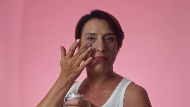 Head shot of mature woman applying anti-age facial cream and looking at camera in pink background, copy space