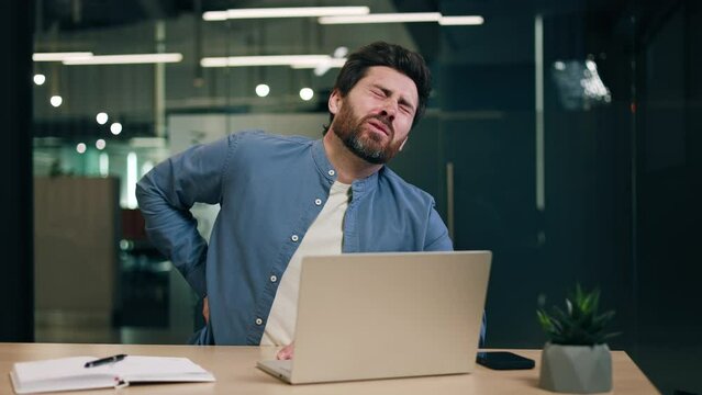 Tired freelancer sitting by table with laptop and notebook while rubbing back with suffering facial expression. Distant smm manager having office syndrome and main symptom of unhealthy posture.