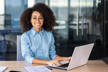 Cheerful diverse woman smiling at camera while keeping hands on portable computer keyboard in...