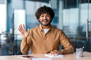 Smiling hispanic employee making hello gesture while keeping hand on table with daybook, pen and tablet. Pleasant bearded man welcoming colleague in meeting room before beginning of discussion.