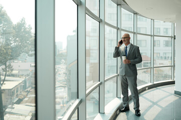 Confident mature chief executive officer with smartphone by ear and cup of coffee in hand standing...