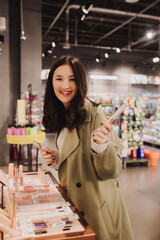 Beautiful woman testing and buying cosmetics in a beauty store. Young girl trying lipstick on hand at cosmetic store. Retail and consumerism. Personal care and modern cosmetology. Side view