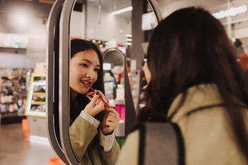 Beautiful teenage testing and buying cosmetics in a beauty store. Young girl trying lipstick on hand at cosmetic store. Retail and consumerism. Personal care and modern cosmetology. Side view