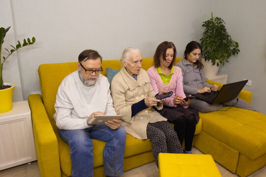 Family members use a laptop, tablet PC and smartphones.Grandfather, grandmother, middle-aged woman and young woman are sitting on the sofa. They use various modern electronic portable devices.