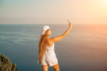 Selfie woman sea. The picture depicts a woman in a cap and tank top, taking a selfie shot with her mobile phone, showcasing her happy and carefree vacation mood against the beautiful sea background