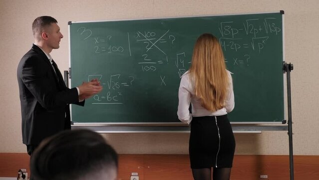 A young female student stands near a blackboard with mathematical formulas written on it. The teacher is standing next to the student. The dialogue between the teacher and the student.