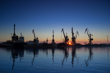 Sunrise at seaport, silhouettes of cranes reflect in water, beginning of cargo operations, supply chain logistics, import export industry, maritime transport, dawn breaks over harbor scenery.