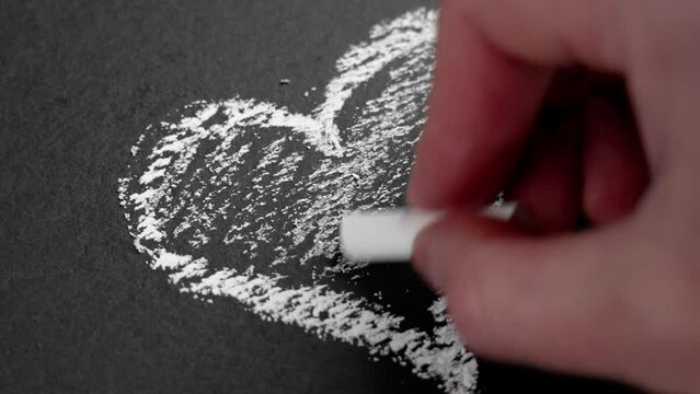 A hand drawing a heart in chalk and hatching a romantic symbol on a black stone surface. Creativity and love concept