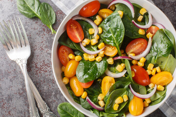 Dietary vegetarian salad of baby spinach, corn, cherry tomatoes and onions dressed with olive oil close-up in a bowl on the table. Horizontal top view from above