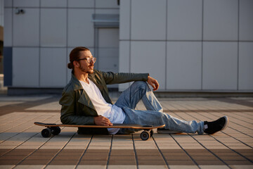 Fashioned hipster guy rest with skateboard sitting in asphalt street road