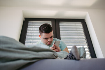 Young man with down syndrome lying in bed, looking at smartphone in morning. Morning routine for...