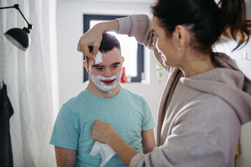 Mother applying shaving foam on son's face. Young man with down syndrome learning how to shave.