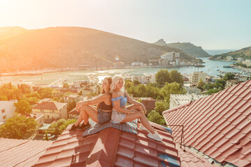 Two women sitting on a red roof, enjoying the view of the town and the sea. Rooftop vantage point....