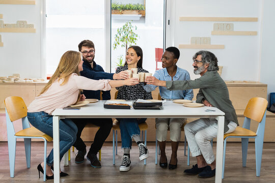 Smiling multiracial group of young people having breakfast, laughing and cheering drinks together at office - Diverse millennial colleagues enjoying meal during work break