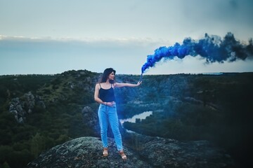 person on the top of the mountain. A Woman Expresses Her Freedom and Imagination with Blue Smoke in Nature. The Evening Sky and the Rocky Landscape Create a Dramatic Contrast.