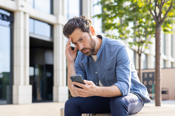 An upset young man sits on a bench on a city street and looks disappointedly at the screen of the phone he is holding in his hands