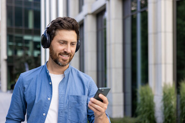 Close-up photo of a young smiling man standing on a city street wearing headphones and using a...