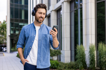 A smiling young man is walking in the city on the street, wearing a denim shirt and his hand in his pants, wearing headphones and using a mobile phone