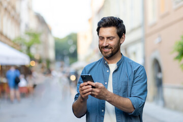 A smiling young man is standing on a city street in a denim shirt and using a mobile phone. Close-up photo
