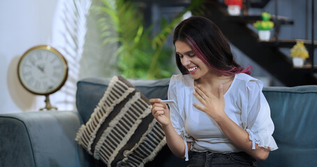Indian Asian woman sitting on sofa couch hold test stick kit look surprise result enjoy day fun joy...