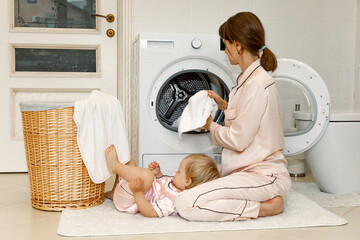 Cute little daughter and family mother in pink pajamas washing dirty clothes in washer in laundry room at home. washing and cleaning concept. daily routine