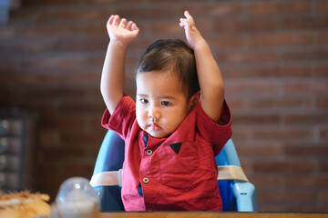 A baby boy sitting in a baby high chair is raising his hands; serious expression