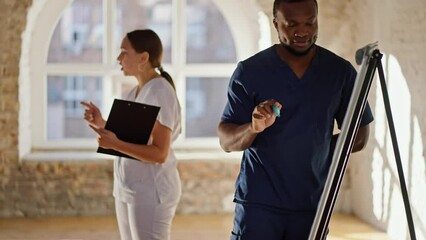 Professional nurse girl in white medical uniform leads medical training during Black man in blue medical uniform records information on canvas with felt-tip pen