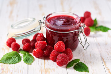 Glass jar of raspberry jam with fresh berries on a white wooden table.