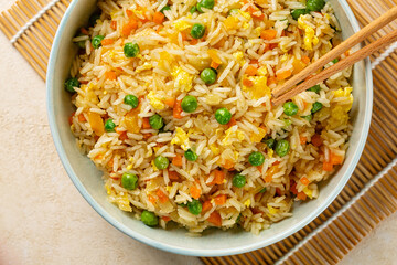 Top view of a bowl with fried rice with egg and vegetables, onion, bell pepper, carrot, green peas. Prepared and served in a light blue plate on a beige table.