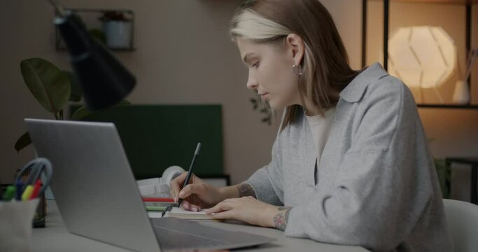Slow motion portrait of young businesswoman working from home using laptop and taking notes at desk at home in evening. Business and remote job concept.