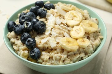 Tasty oatmeal with banana, blueberries, coconut flakes and honey served in bowl on beige table, closeup