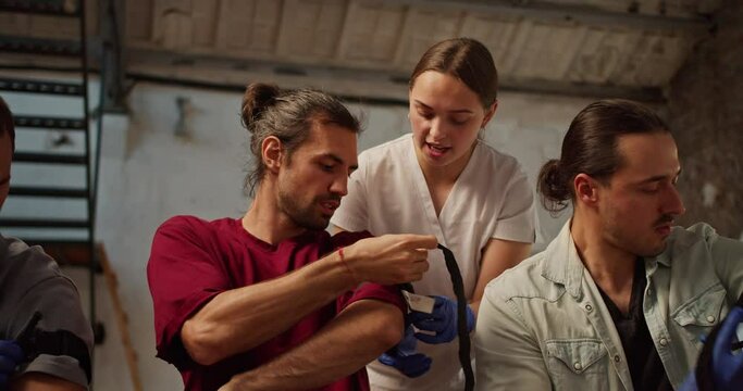 Close-up shooting: A professional nurse in a white medical uniform helps a brunette man with long hair to properly put a tourniquet on his shoulder during a practical training in medicine