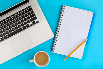 Blank notebook, laptop and cup of coffee on a blue background, top view.