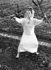 Beautiful woman in a dress dances barefoot on a muddy field, holding reeds in her hands