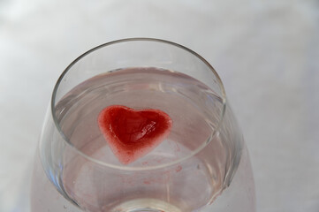 A glass is full of a clear liquid and a bright red, heart shaped ice cube floating in it