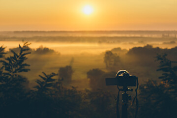 Silhouette of a camera on a tripod on a hill capturing sunrise and morning fog. Countryside landscape with camera on tripod, sunrise and morning fog.
