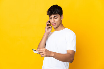 Young man over isolated yellow background buying with the mobile with a credit card