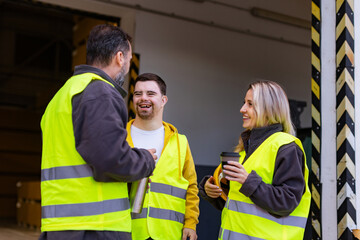 Man with Down syndrome and his colleagues taking break from work, drinking coffee, hot tea outdoors, on loading dock. Concept of workers with disabilities, support in workplace.