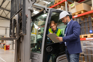 Female forklift driver talking with manager in warehouse, order picking. Warehouse worker preparing products for shipmennt, delivery, checking stock in warehouse.