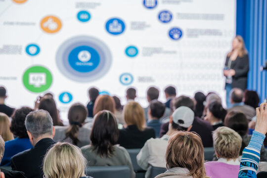 Focused audience listening to a female speaker at a business conference with informational graphics.