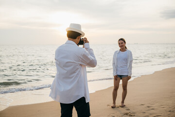Romantic sunrise at beach for young couple in white as boyfriend takes candid photos of his...