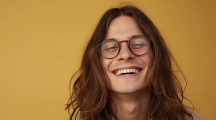 Fototapeta na wymiar Young man with long brown hair wearing round glasses smiling broadly against a yellow background.