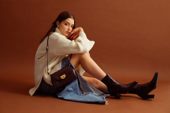 Fashionable confident woman wearing white sweater, denim skirt, pointed toe ankle leather boots, with trendy shoulder bag, posing on brown background. Full-length studio fashion portrait. Copy space