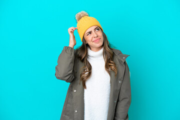 Young Italian woman wearing winter jacket and hat isolated on blue background having doubts and...